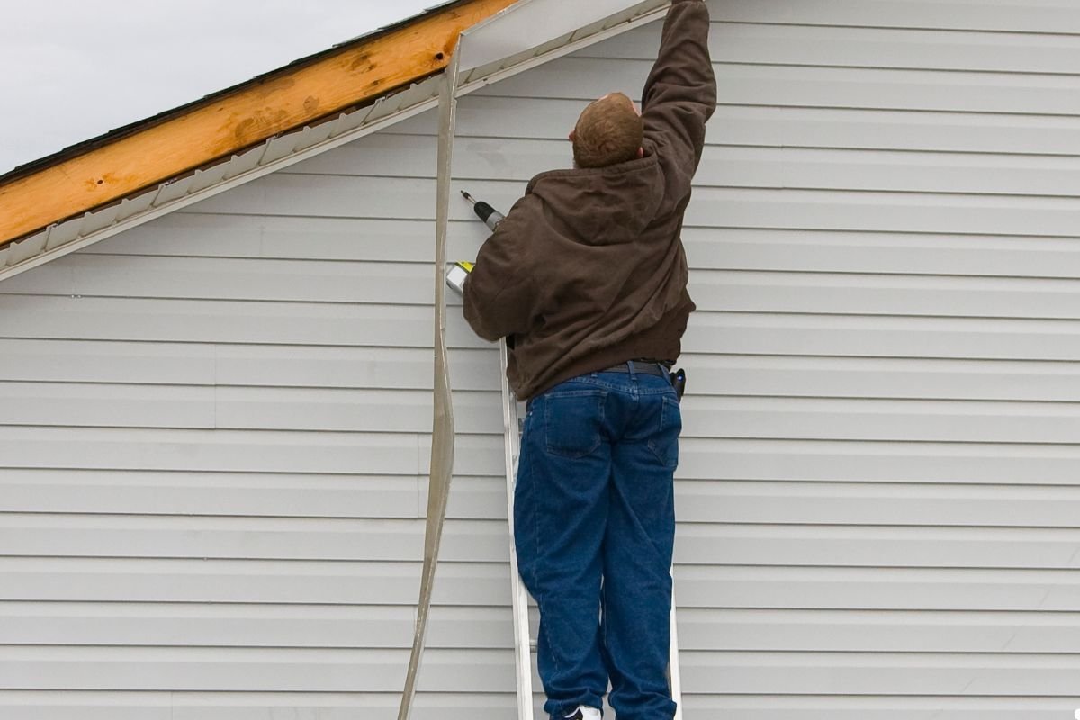 A man stands on a ladder, painting the exterior siding of a house with a brush, surrounded by tools and paint cans.