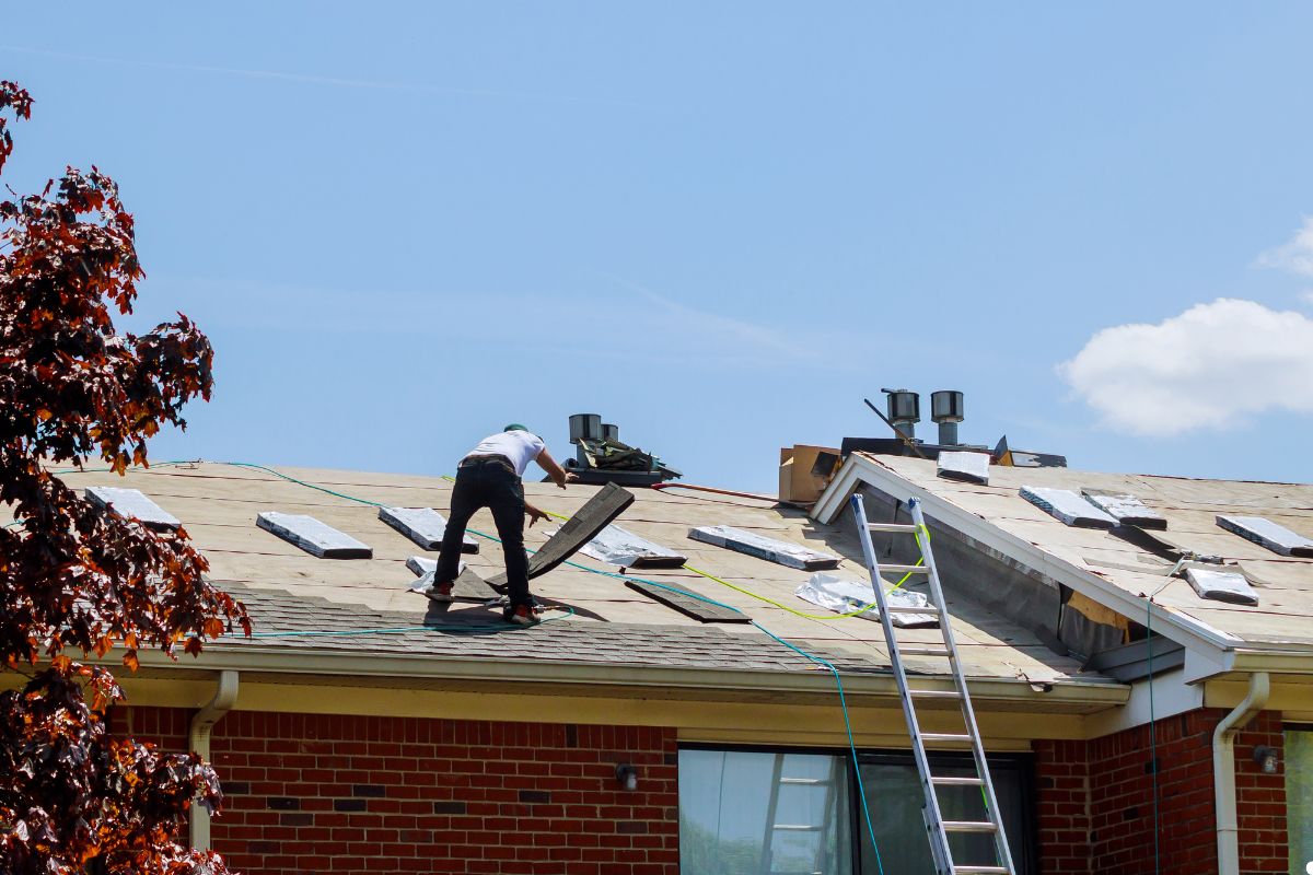 A man is repairing the roof of a house, wearing safety gear and focused on his work under a clear blue sky.
